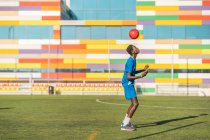 African American teenager in blue uniform bouncing bright ball on head during training on football field — Stock Photo