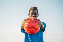 Черный подросток в синей футболке с красным мячом в вытянутых руках и смотрящий в камеру на безоблачное небо — стоковое фото
