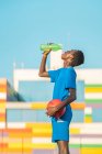 African American teenager with ball drinking fresh water during football training on sunny daytime — Stock Photo