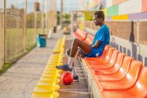 Vista lateral do jogador de futebol afro-americano descansando em assentos de estádio — Fotografia de Stock