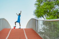 Schwarzer Teenager in blauer Sportbekleidung tritt beim Training auf der Straße auf den Fußballball und hebt die Arme — Stockfoto