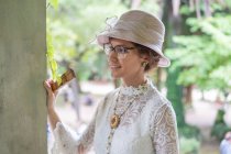 Элегантная женщина в ретро шляпе и кружевной блузке улыбается и смотрит в сторону, стоя возле колонны на размытом фоне парка — стоковое фото