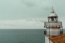 From above white lighthouse stayed next to building with red tiled roof on sea shore with calm dark water and grey cloudy sky — Stock Photo
