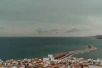 Вид с воздуха на морской порт с плотно стоящими зданиями с красными крышами и пирсом с лодками на побережье с темной водой и серым облачным небом — стоковое фото