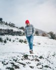 Випадковий чоловік у червоній в'язаній шапці в смугастому светрі та джинсах, що ходять на зимовому полі з пагорбами, покритими снігом — стокове фото