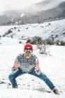 Casual man in red knitted cap playing with snow on winter field with hills — Stock Photo