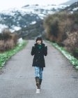 Jolie jeune femme en manteau noir et jean marchant sur une route de campagne vide avec des collines couvertes de neige — Photo de stock