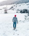 Visão traseira do macho casual em gorro de malha vermelho andando no campo de inverno com colinas cobertas de neve — Fotografia de Stock
