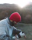 Вид збоку бородатого чоловіка-фотографа в червоному в'язаному в'язанні захоплює професійну камеру в рюкзаку в горах — стокове фото