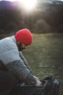 Seitenansicht eines bärtigen männlichen Fotografen mit roter Strickmütze, der in den Bergen nach einer professionellen Kamera im Rucksack greift — Stockfoto
