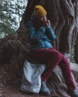 Junges Mädchen in warmer, aktiver Kleidung sitzt mit Rucksack auf alten massiven Baumwurzeln und wärmenden Händen — Stockfoto