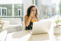 Етнічна жінка з ноутбуком насолоджується здоровим напоєм у кафе — стокове фото