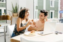 Cheerful ethnic woman enjoying fresh beverage and leaning on shoulder of boyfriend with laptop while sitting at cafe table — Stock Photo