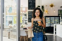 Mujer étnica feliz con teléfono inteligente y jugo fresco sonriendo y mirando hacia otro lado mientras deja la cafetería moderna - foto de stock