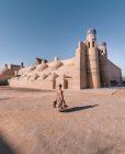 Visão traseira do turista adulto em vestido colorido de pé sozinho na rua contra edifícios antigos marrons e torres com paredes de adobe e cúpulas em Khiva durante o tempo justo — Fotografia de Stock