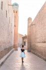 Visão traseira da fêmea em saia romântica em pé sozinho na rua estreita contra edifícios antigos marrons com paredes de adobe no Khiva Uzbequistão — Fotografia de Stock