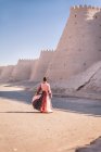 Back view of female in colorful traditional dress spinning around against ancient high winding stone defensive wall around inner town in Khiva under clear blue sky — Stock Photo