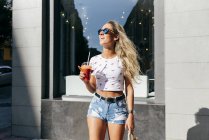 Fröhliche Frau im trendigen Outfit mit frischem Getränk schaut weg, während sie auf der Stadtstraße steht — Stockfoto