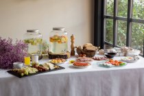 Vista lateral do jantar festivo com saborosa comida de padaria de frutas e elegantes margens de limonada na mesa decorada com flores lilás — Fotografia de Stock