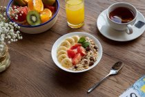 Сверху чаша гранолы с вкусными ягодами и бананом, подаваемая на стол с тарелкой фруктов и чашкой чая с завтраком — стоковое фото