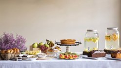 Vista lateral de la cena festiva con sabrosa comida de panadería de frutas y elegantes bancos de limonada en la mesa decorada con flores de lila. - foto de stock