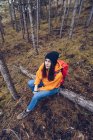 From above view of woman in warm clothes with backpack sitting on log and looking in camera in evergreen forest — Stock Photo