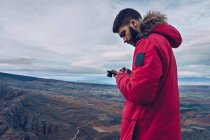 Вид сбоку внимательного мужчины в куртке, пользующегося смартфоном, стоя на скале над захватывающим дух видом — стоковое фото