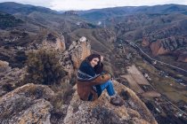 Von oben positive Frau blickt in die Kamera und sitzt auf Felsen am Rande der Klippe mit malerischer Aussicht — Stockfoto