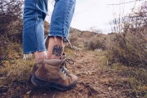 Beine tätowierter Wanderer in Stiefeln und Jeans, der auf schlammigem Pfad am Herbstfeld mit getrocknetem Gras spaziert — Stockfoto