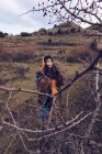 Giovane donna in abiti caldi in piedi sul campo con foglie secche da alberi nudi e guardando in macchina fotografica — Foto stock