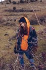 Giovane donna in abiti caldi in piedi sul campo con foglie secche da alberi nudi e guardando in macchina fotografica — Foto stock