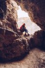 Беззаботная женщина в теплой одежде сидит у входа в скалистую дыру при дневном свете — стоковое фото