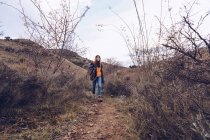 Jovem com mochila brilhante em roupas quentes andando ao longo da estrada com folhas secas por árvores nuas — Fotografia de Stock