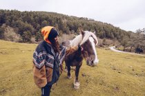 Любящая женщина в теплой одежде гладит спокойную лошадь, стоя на деревенском лугу у леса — стоковое фото