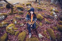 From above woman in hat sitting in stones surrounded with dried foliage and moss and looking in camera — Stock Photo
