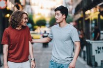Despreocupados hombres multiétnicos interesados en ropa casual hablando mientras pasean por la calle de la ciudad - foto de stock