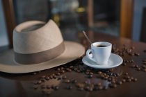 Coupe en céramique avec cuillère à café parmi les grains de café torréfiés à côté du chapeau sur la table en bois — Photo de stock