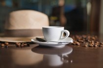 Керамічна чашка з чайною ложкою серед обсмажених кавових зерен біля капелюха на дерев'яному столі — стокове фото