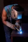 Вид збоку майстра в захисній масці для обличчя в комбінезоні та рукавичках, що працюють в майстерні та паяльнику металу — стокове фото
