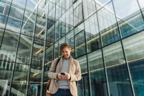 Low angle of happy businessman using smartphone while standing outside business center on city street — Stock Photo
