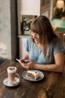 Блондинка весела жінка друкує повідомлення на смартфоні, сидячи за столом з кавою і десертом у затишному кафе — стокове фото