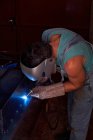 Ремесленник в защитной маске для лица в комбинезонах и перчатках, работающих в мастерской и паяльной металлической конструкции — стоковое фото