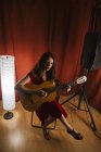 Charmante künstlerische Frau im roten Kleid, die bei warmem Licht auf der Bühne ein Lied auf der Gitarre spielt — Stockfoto