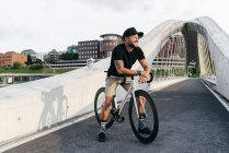 Happy adult bearded man in black cap wearing black shirt and beige shorts sitting resting on bicycle across footbridge in city — Stock Photo