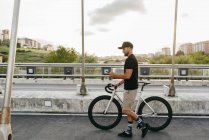 Side view of young casual bearded male cyclist in black cap using phone while walking with bike on road with city buildings on background — Stock Photo