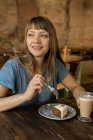 Блондинка весела щаслива жінка з чубчиком тримає ложку з шматочком торта і сидить за столом з кавою і десертом — стокове фото