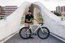 Homem barbudo adulto feliz em boné preto vestindo camisa preta e shorts bege de pé com bicicleta através da ponte na cidade olhando para a câmera — Fotografia de Stock