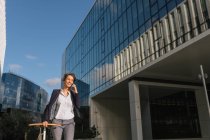 Positive Geschäftsfrau mit Fahrrad lächelt und spricht auf dem Smartphone, während sie vor einem modernen Gebäude spaziert — Stockfoto