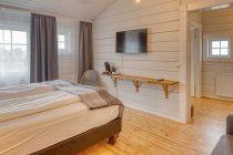 Cómodo dormitorio con paredes de madera blanca y cama grande y suave con acogedor sofá gris cerca de la lámpara de pie en llamas - foto de stock