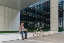 Happy man manager smiling and browsing laptop while sitting outside near bicycle on a contemporary building on city street — Stock Photo